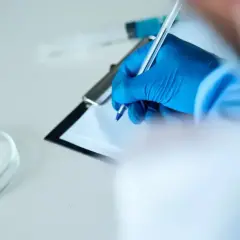 Ręka w niebieskiej rękawiczce pisze na kartce. Obok szalka Petriego z białym proszkiem i strzykawka. To może być test na narkotyki.