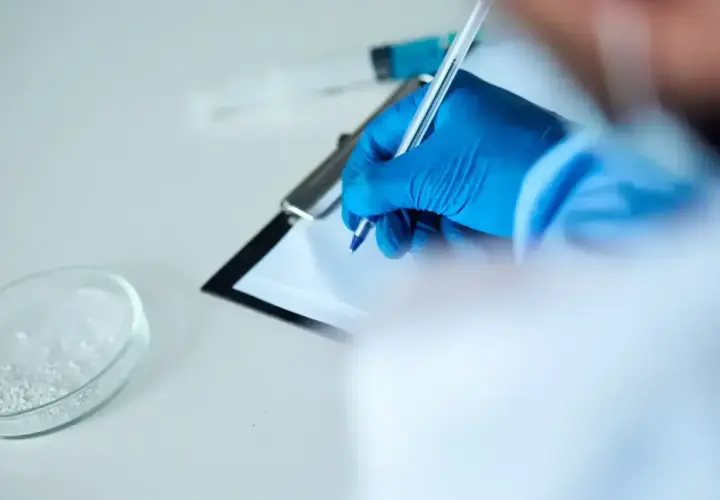 Ręka w niebieskiej rękawiczce pisze na kartce. Obok szalka Petriego z białym proszkiem i strzykawka. To może być test na narkotyki.