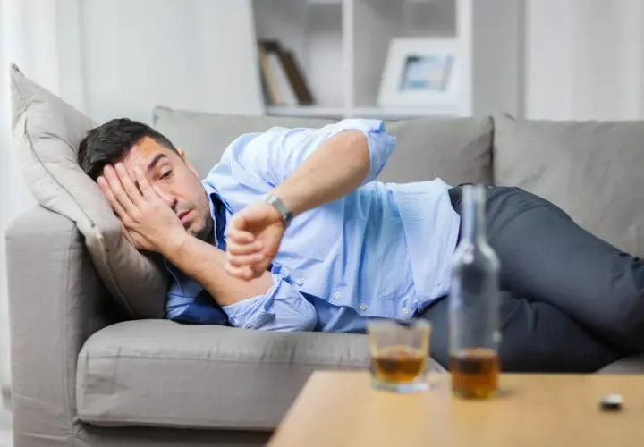 Mężczyzna leży na kanapie, zasłaniając twarz ręką, obok butelka i szklanka alkoholu. Zastanawia się, jak szybko wytrzeźwieć.