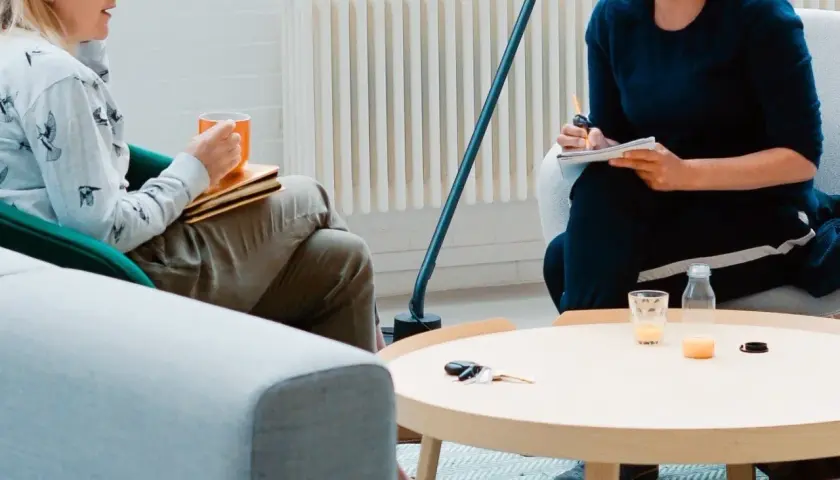 Dwie osoby siedzą w fotelach, jedna trzyma kubek, druga pisze w notatniku.