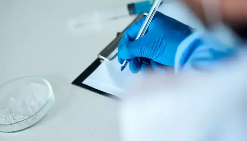 Ręka w niebieskiej rękawiczce pisze na kartce. Obok szalka Petriego z białym proszkiem i strzykawka. To może być test na narkotyki.