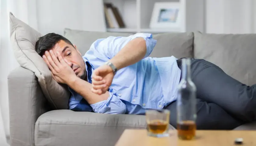 Mężczyzna leży na kanapie, zasłaniając twarz ręką, obok butelka i szklanka alkoholu. Zastanawia się, jak szybko wytrzeźwieć.