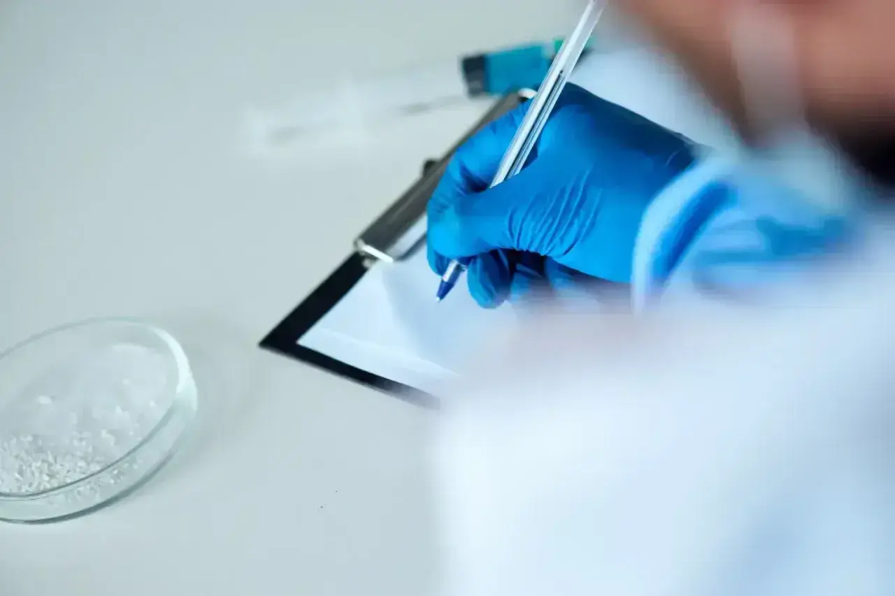 Ręka w niebieskiej rękawiczce pisze na kartce. Obok szalka Petriego z białym proszkiem i strzykawka. To może być test na narkotyki.